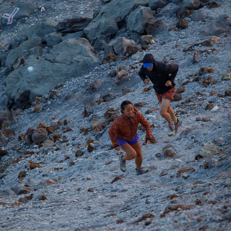 Two trail runners ascending a rugged mountain slope with loose rocks and boulders, one in a bright orange jacket and blue shorts, the other in black, against a twilight sky.