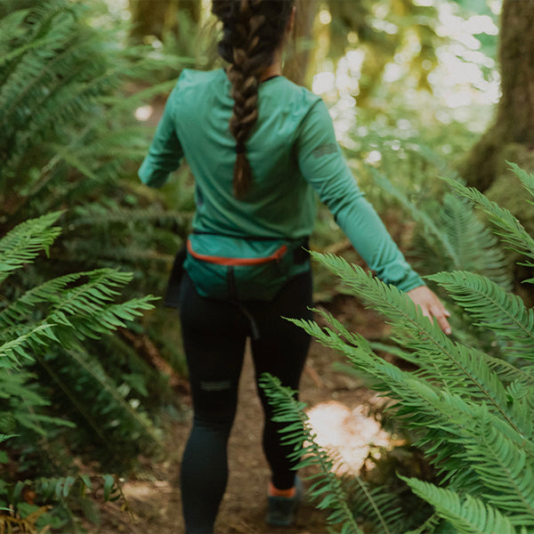 A woman with a braided ponytail wearing a teal long-sleeve shirt and black leggings running on a forest trail, with ferns and dense woodland surrounding her.