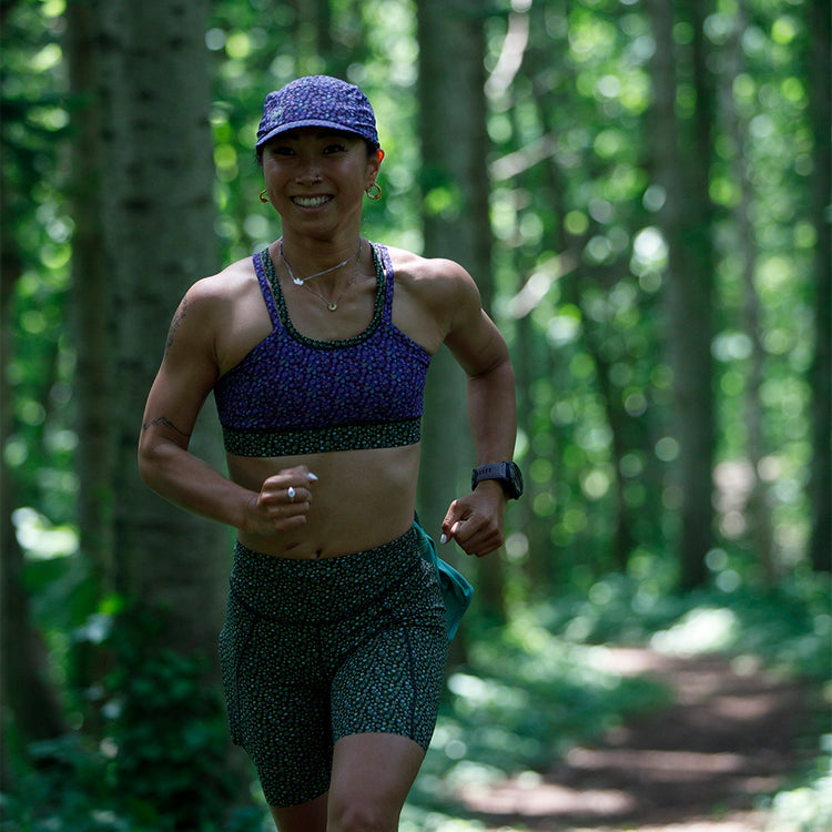 A smiling runner with a matching patterned sports bra and shorts set, accessorized with a cap and a wristwatch, jogging through a sun-dappled forest trail.