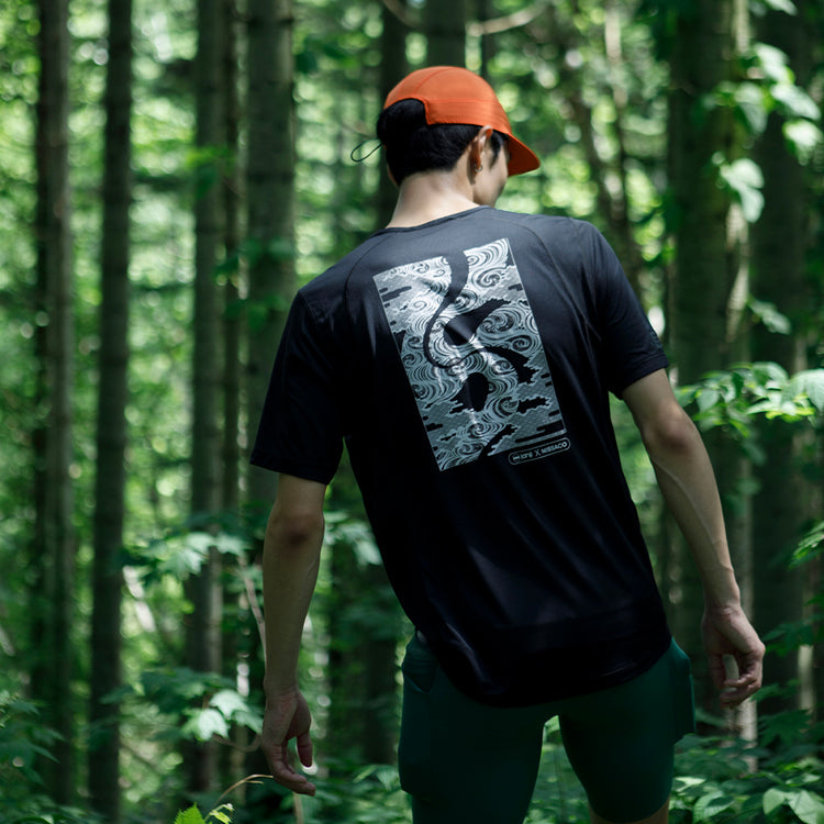 A person from behind, wearing a black t-shirt with a distinctive white graphic design and a red cap, walking through a forest with tall, slender trees.