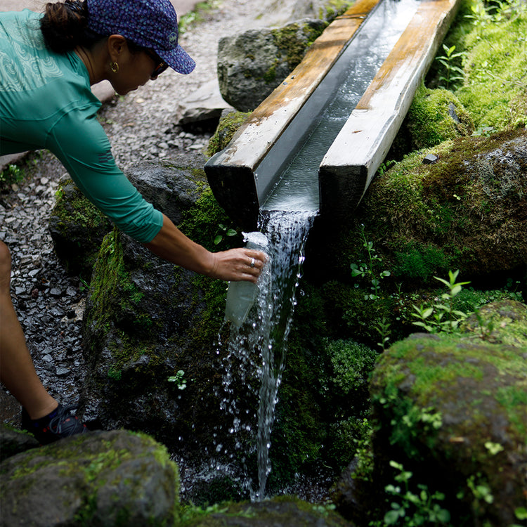 A runner refilling a water bottle from a clear mountain stream flowing through a wooden aqueduct, surrounded by lush greenery and moss-covered rocks.